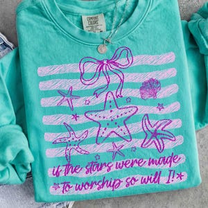 Op de afbeelding: Teal Comfort Colors sweatshirt met een roze zeester en strik ontwerp. Het ontwerp bevat horizontale strepen, zeesterren en de tekst "if the stars were made to worship so will I!" Een zilveren ketting is zichtbaar.