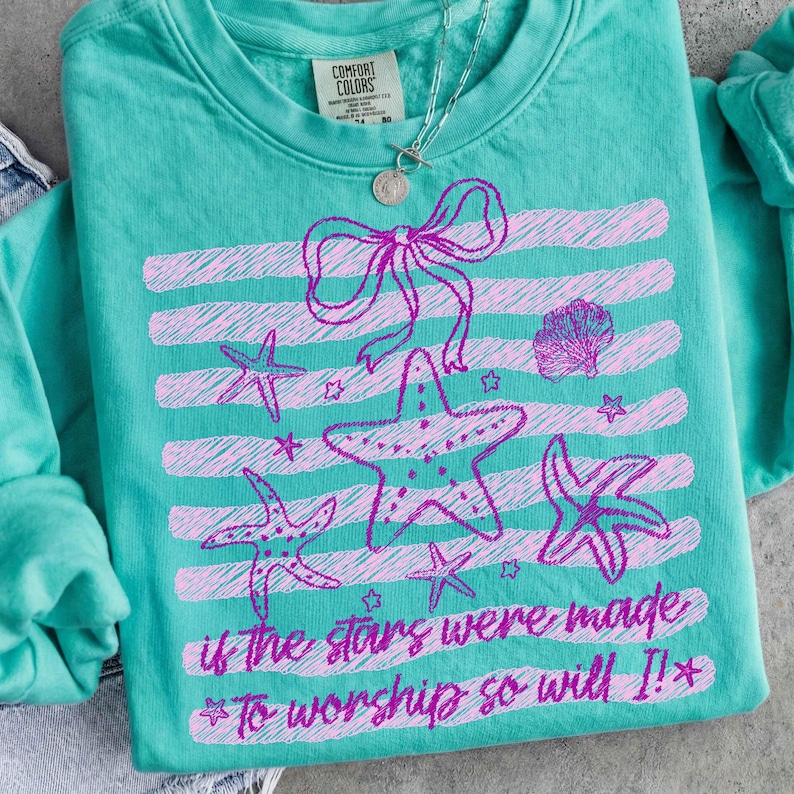 Op de afbeelding: Teal Comfort Colors sweatshirt met een roze zeester en strik ontwerp. Het ontwerp bevat horizontale strepen, zeesterren en de tekst "if the stars were made to worship so will I!" Een zilveren ketting is zichtbaar.