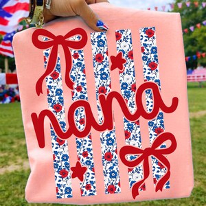 Op de afbeelding: Een lichtroze t-shirt met het woord "nana" in rood script. Het ontwerp bevat rode strikken en sterren, samen met verticale strepen gevuld met een bloemenpatroon in rood, wit en blauw. Het shirt wordt door een hand omhoog gehouden.