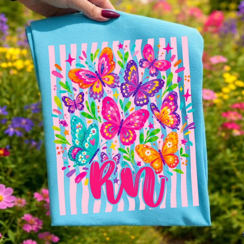 May include: A light blue t-shirt featuring a graphic print of colorful butterflies in shades of pink, orange, purple, and teal. The design includes vertical light pink stripes, small green leaves, and the stylized pink text RN at the bottom.
