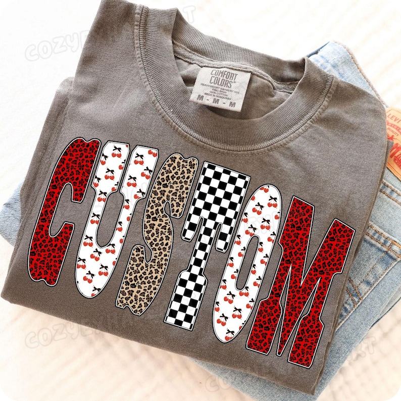 May include: A gray Comfort Colors t-shirt with the word "CUSTOM" in a bold, decorative font. The letters feature cherry, leopard, and checkered patterns. The shirt is folded on top of a pair of blue jeans.