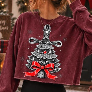 K&ouml;nnte beinhalten: Ein bordeauxrotes, lang&auml;rmeliges bauchfreies Sweatshirt mit einer Grafik eines stilisierten Weihnachtsbaums. Der Baum ist mit einer gro&szlig;en roten Schleife an der Basis, einem Stern an der Spitze und kleinen Kugeln verziert, umgeben von wei&szlig;en Schneeflocken.