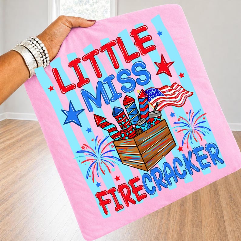 May include: A pink blanket with blue stripes and the words "LITTLE MISS FIRECRACKER" in red and blue. The blanket features a graphic of fireworks, stars, and an American flag.