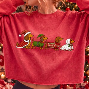 May include: A red long-sleeve sweatshirt featuring a graphic of four dachshunds dressed in holiday attire. The dogs are wearing a Santa hat and cape, an elf outfit, a red patterned knit jumper, and a white hooded cloak with holly accents.