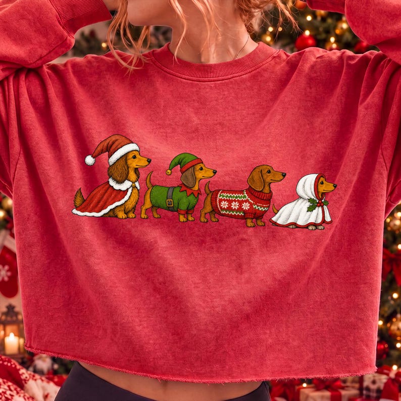 May include: A red long-sleeve sweatshirt featuring a graphic of four dachshunds dressed in holiday attire. The dogs are wearing a Santa hat and cape, an elf outfit, a red patterned knit jumper, and a white hooded cloak with holly accents.