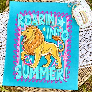 May include: A turquoise towel with a cartoon lion illustration and the words "ROARIN' INTO SUMMER!" in pink and blue. The lion is yellow with an orange mane and tail. The design includes stars and a wavy border.