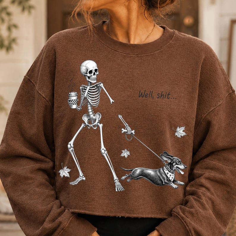 May include: A brown long-sleeve sweatshirt featuring a graphic of a skeleton holding a coffee cup and walking a dachshund on a leash. The text "Well, shit..." is printed above the dog, accompanied by small illustrations of falling autumn leaves.