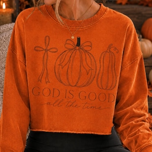 K&ouml;nnte beinhalten: Ein lang&auml;rmeliges Sweatshirt in gebranntem Orange mit einem Saum mit Rohkante. Die Vorderseite zeigt eine schwarze Strichzeichnung eines Kreuzes mit Schleife, eines K&uuml;rbisses mit Schleife und eines K&uuml;rbisgew&auml;chses. Der Text lautet "GOD IS GOOD all the time".