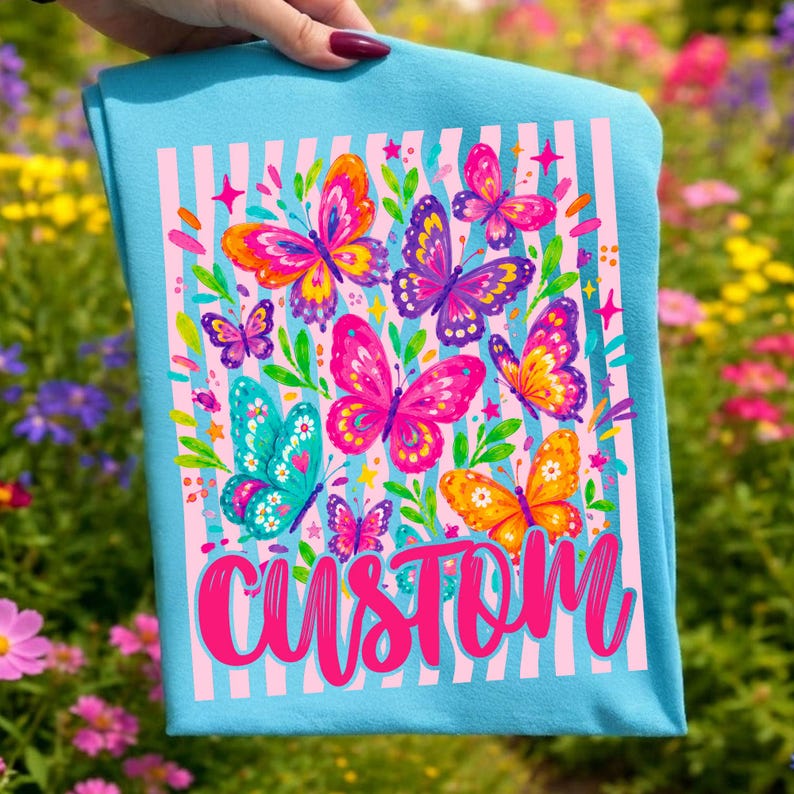 May include: A light blue t-shirt featuring a graphic print of colourful butterflies in shades of pink, orange, purple, and teal. The design includes vertical light pink stripes, small green leaves, and the word CUSTOM written in bold pink script.
