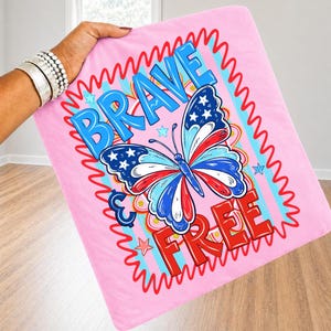 May include: Pink blanket featuring a butterfly design in red, white, and blue, with the words "BRAVE & FREE". The butterfly wings have stars and stripes. The blanket is held by a person.