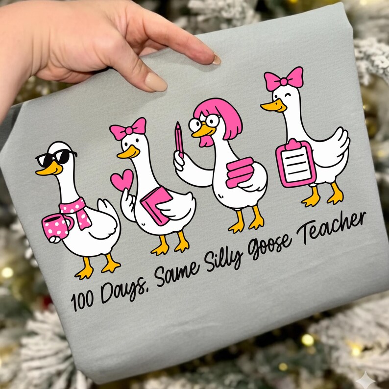 K&ouml;nnte beinhalten: Ein hellgraues T-Shirt mit einem Cartoon-Design von vier G&auml;nsen, jede mit einzigartigen Accessoires wie Sonnenbrillen, Schleifen und B&uuml;chern. Der Text "100 Days, Same Silly Goose Teacher" ist darunter gedruckt.