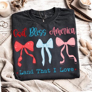 Może przedstawiać: Ciemnoszara koszulka z napisem "God Bless America" w kolorze czerwonym i niebieskim oraz "Land That I Love" w kolorze niebieskim. Koszulka posiada trzy kokardy w kolorze czerwonym, jasnoniebieskim i r&oacute;żowym.