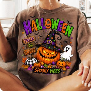 May include: A brown t-shirt featuring a graphic print with the text HALLOWEEN, TRICK OR TREAT, and SPOOKY VIBES. The design includes a large jack-o'-lantern wearing a witch hat, a white ghost, a buffalo plaid pumpkin, and small bats and spiders.
