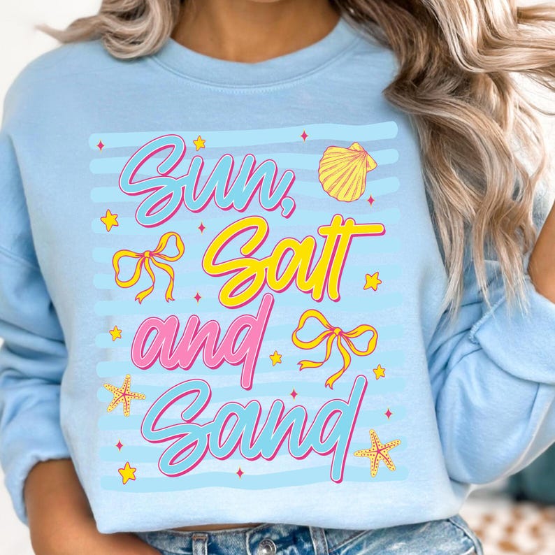 Peut inclure: Sweat-shirt bleu clair avec l'inscription "Sun, Salt and Sand" dans un design color&eacute; sur le th&egrave;me de la plage. Le texte est en rose et jaune, avec des accents de coquillages et d'&eacute;toiles de mer. Le sweat-shirt a des manches longues.