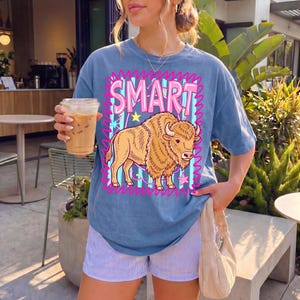 May include: Blue t-shirt with the word "SMART" in pink letters and a cartoon bison illustration. The shirt has a vintage wash look. The person is wearing blue and white striped shorts and holding a coffee drink.