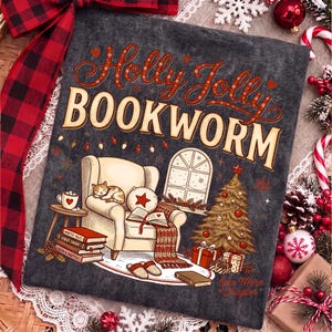 Op de afbeelding: Een donkergrijs t-shirt met de tekst Holly Jolly BOOKWORM in rood en cr&egrave;me. De afbeelding toont een gezellige fauteuil met een slapende kat, een open boek, een kerstboom met cadeautjes, een raam en een stapel boeken met Winter Tales.