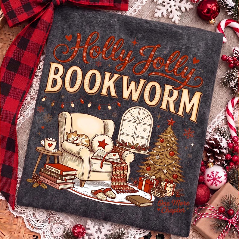 Op de afbeelding: Een donkergrijs t-shirt met de tekst Holly Jolly BOOKWORM in rood en cr&egrave;me. De afbeelding toont een gezellige fauteuil met een slapende kat, een open boek, een kerstboom met cadeautjes, een raam en een stapel boeken met Winter Tales.