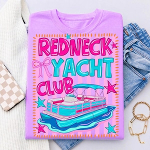 May include: A lilac-colored t-shirt with the words "REDNECK YACHT CLUB" in pink and teal. The design includes a cartoon pontoon boat and stars. The shirt is displayed with a white sweater, jeans, a phone case, and a gold bracelet.
