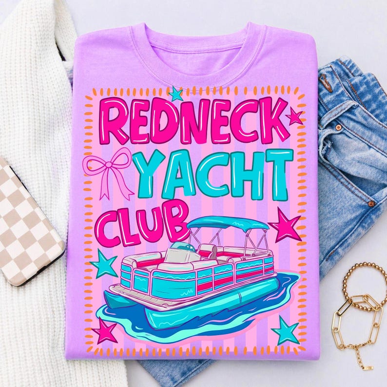 May include: A lilac-colored t-shirt with the words "REDNECK YACHT CLUB" in pink and teal. The design includes a cartoon pontoon boat and stars. The shirt is displayed with a white sweater, jeans, a phone case, and a gold bracelet.