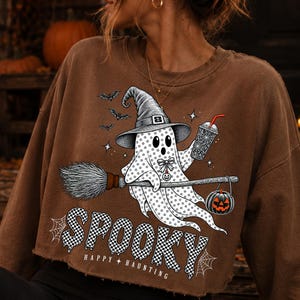 May include: A brown cropped sweatshirt featuring a graphic of a ghost wearing a witch hat, riding a broomstick, and holding a drink. The ghost is decorated with stars and a bow. Below, the text reads SPOOKY and HAPPY HAUNTING in a checkered pattern.