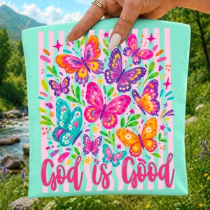 May include: A light mint green tote bag featuring a vertical pink striped pattern and a vibrant collection of colorful butterflies in shades of pink, orange, yellow, and blue. The text God is Good is printed in a bold pink script at the bottom.