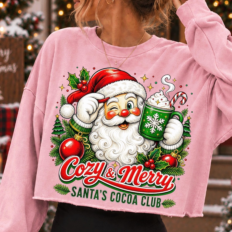 K&ouml;nnte beinhalten: Ein hellrosa bauchfreies Langarm-Sweatshirt mit einer Grafik eines zwinkernden Weihnachtsmanns, der einen gr&uuml;nen Becher mit hei&szlig;er Schokolade und einer Zuckerstange h&auml;lt. Der Text lautet Cozy & Merry SANTA'S COCOA CLUB, umgeben von Stechpalmen und Tannen.
