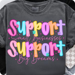 May include: Dark gray sweatshirt with the words "Support Small Businesses Support Big Dreams" in colorful, bubble letters. The text is decorated with small flower graphics. The sweatshirt is a casual style.