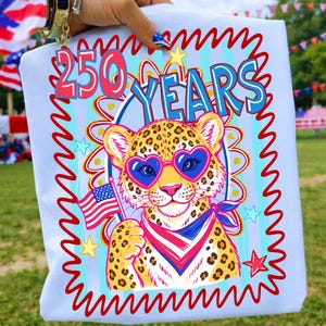 Op de afbeelding: Lichtblauw t-shirt met een cartoon luipaard met een hartvormige zonnebril en een patriottische bandana, met een kleine Amerikaanse vlag. Het shirt heeft de tekst "250 YEARS" in rood en blauw, met een rode en blauwe golvende rand.