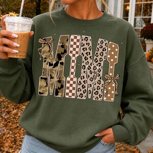 May include: An olive green crewneck sweatshirt featuring the word MIMI in large block letters. Each letter has a unique pattern: camouflage, brown and white checkerboard, leopard print, and tan with white polka dots, accented by two brown bows.