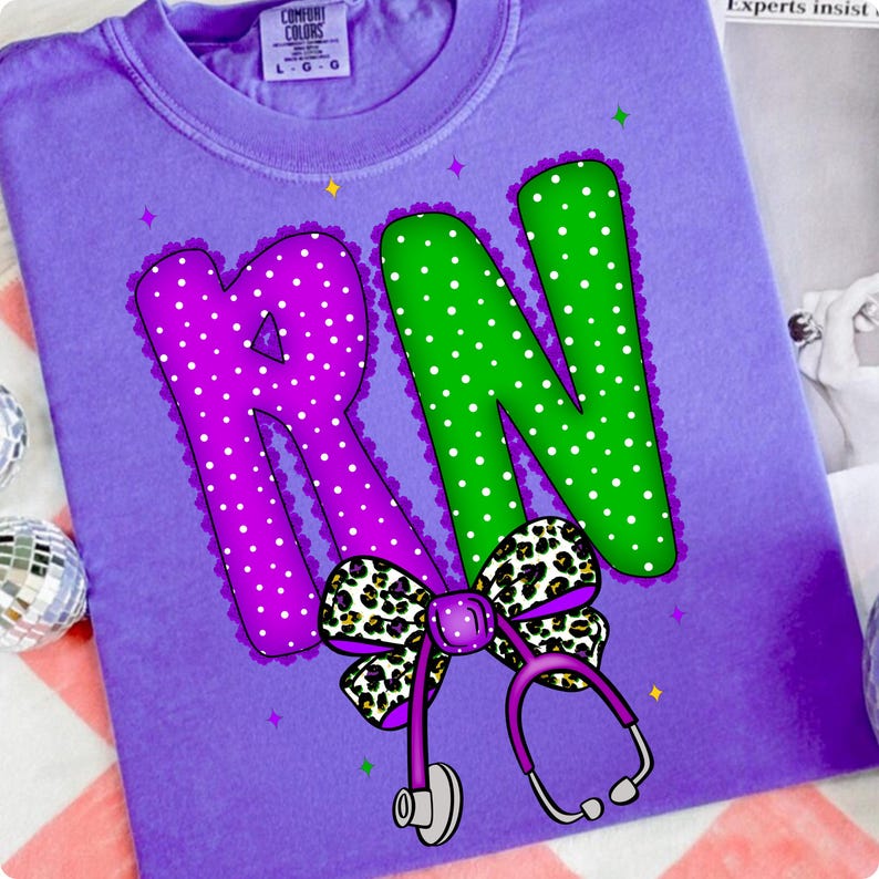May include: A purple t-shirt with a graphic design featuring the letters "RN" in purple and green with white polka dots. A leopard print bow and a purple stethoscope complete the design. The shirt is from the brand Comfort Colors.