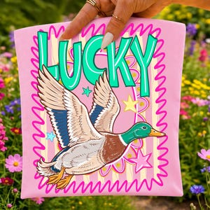 May include: A pink t-shirt with a cartoon duck in flight, and the word "LUCKY" in teal letters. The design includes stars and a wavy pink border. The duck has brown, blue, and white feathers.