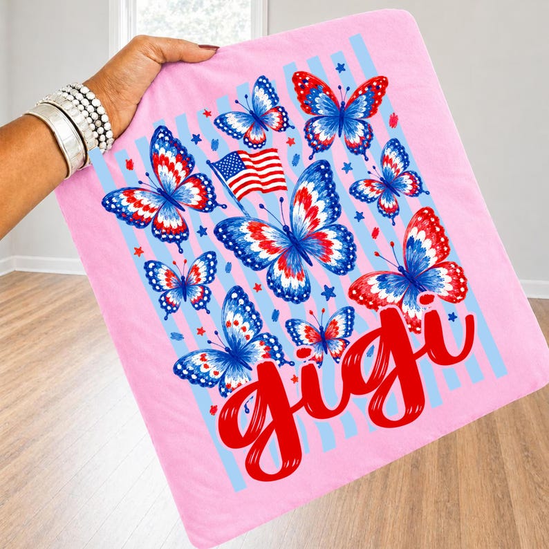May include: A light pink square fabric item featuring a graphic print of multiple butterflies with red, white, and blue wing patterns. The design includes a small American flag, scattered blue stars, light blue vertical stripes, and the word gigi in red.