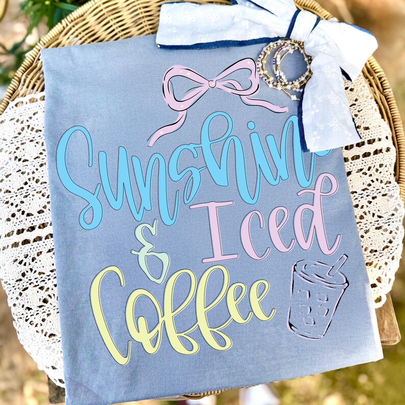 Op de afbeelding: Een lichtgrijs t-shirt met de woorden "Sunshine & Iced Coffee" in pastelkleuren. Een afbeelding van een ijskoffiebeker staat ook op het shirt. Een witte strik met denim afwerking en een roze strik staan bovenaan.