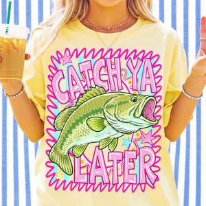 Op de afbeelding: Geel T-shirt met een afbeelding van een groen en gele vis met open mond. De tekst "CATCH YA LATER" is roze, omlijnd met een golvend blauw en roze ontwerp. Het shirt heeft een casual stijl.