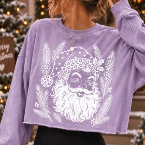 Puede incluir: Una sudadera corta de manga larga en color morado claro con un estampado gr&aacute;fico blanco de un Santa Claus gui&ntilde;ando un ojo, con un gorro de estampado de leopardo, rodeado de ramas de pino y bayas de acebo. La prenda tiene un dobladillo sin rematar.