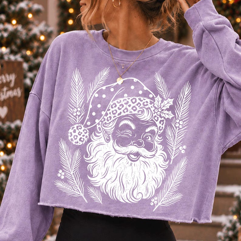 Puede incluir: Una sudadera corta de manga larga en color morado claro con un estampado gr&aacute;fico blanco de un Santa Claus gui&ntilde;ando un ojo, con un gorro de estampado de leopardo, rodeado de ramas de pino y bayas de acebo. La prenda tiene un dobladillo sin rematar.