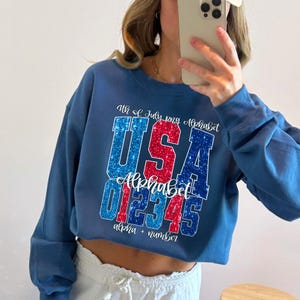K&ouml;nnte beinhalten: Ein blaues Sweatshirt mit einem patriotischen Design. Das Design zeigt das Wort "USA" in rotem und blauem Glitzer, zusammen mit den Zahlen "012345" und den W&ouml;rtern "Alphabet" und "alpha + number". Der Text "4th of July png Alphabet" ist ebenfalls enthalten.