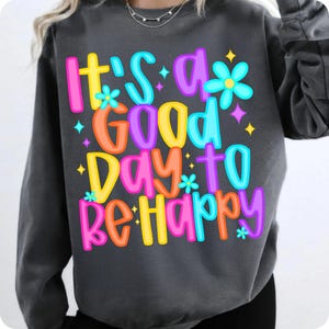 K&ouml;nnte beinhalten: Ein grauer Pullover mit dem Text "It's a good day to be happy" in bunten Buchstaben und Blumen.