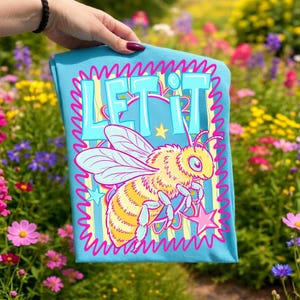 May include: A light blue t-shirt featuring a cartoon bee holding a star-shaped wand. The shirt has the words "LET IT" in a turquoise font, outlined in pink. The bee is yellow and has pink accents. The shirt is held up in front of a colorful flower garden.