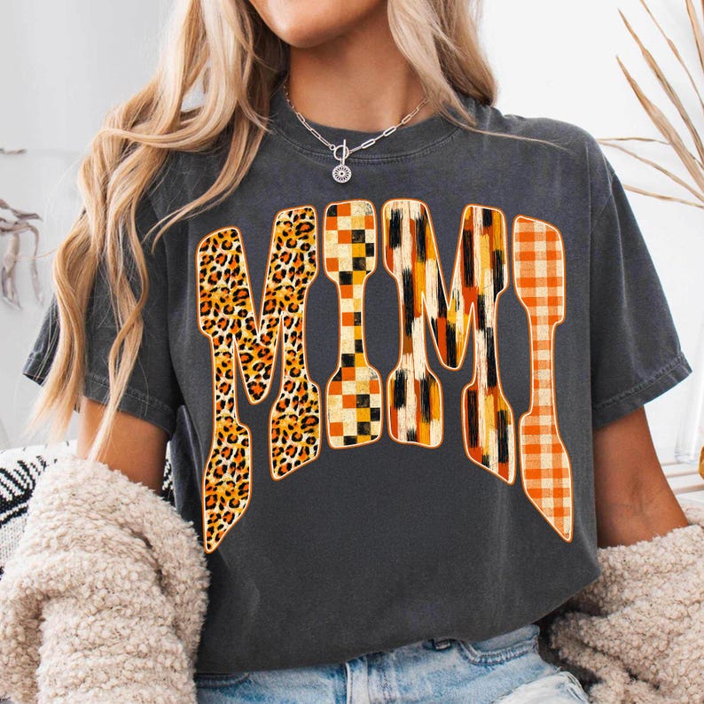 May include: A charcoal gray short-sleeve t-shirt featuring the word MIMI printed in large, bold letters across the chest. Each letter displays a distinct pattern, including leopard print, a black and orange checkerboard, vertical brushstrokes, and orange plaid.