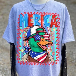 May include: A light blue t-shirt featuring a cartoon duck wearing sunglasses, a hat, and a bandana, all in patriotic colors. The word "MERICA" is printed across the top in red and blue. The design is framed by a red, wavy border.