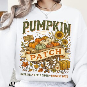 Puede incluir: Una sudadera blanca con un gr&aacute;fico de un carro de madera lleno de calabazas, girasoles y un letrero que dice FRESH PICKED. El texto PUMPKIN PATCH aparece en una cinta, con las palabras HAYRIDES, APPLE CIDER y HARVEST DAYS debajo.
