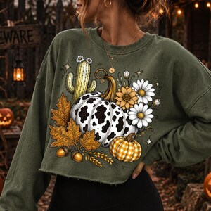 Puede incluir: Una sudadera corta de manga larga en color verde oliva con un estampado gr&aacute;fico de una calabaza con manchas de vaca, una calabaza de cuadros amarillos, un cactus, hojas de arce oto&ntilde;ales, bellotas y margaritas blancas.