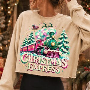 May include: A beige long-sleeve cropped sweatshirt featuring a graphic print of a green and pink steam train, snow-covered pine trees, and Santa in a sleigh. The text "CHRISTMAS EXPRESS" is printed in large, stylised, multi-coloured lettering.