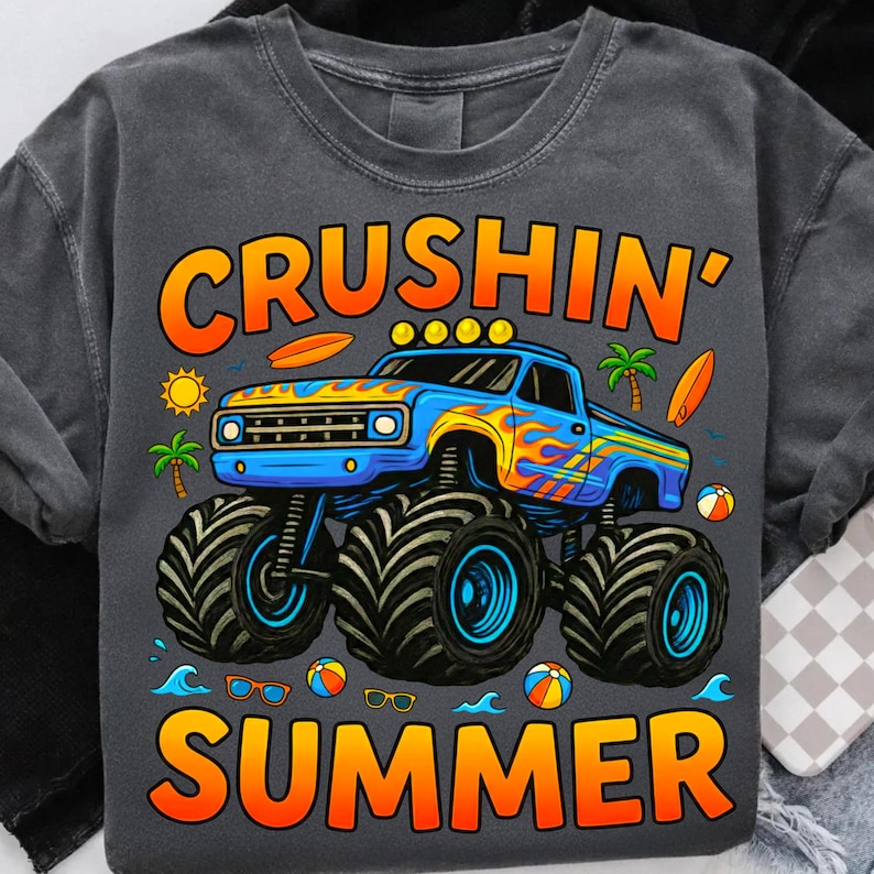 May include: Dark gray t-shirt with a colorful graphic. The graphic features a blue monster truck with flames, the words "CRUSHIN' SUMMER", a sun, palm trees, surfboards, and beach balls. The shirt is a casual summer design.