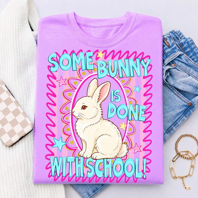 Może przedstawiać: Liliowa koszulka z grafiką białego kr&oacute;lika w owalu. Tekst "SOME BUNNY IS DONE WITH SCHOOL!" jest w kolorze turkusowym i r&oacute;żowym. Projekt zawiera gwiazdki i dekoracyjną obw&oacute;dkę.