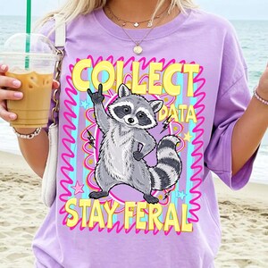 May include: Lilac t-shirt with a cartoon raccoon giving the middle finger. The text "COLLECT DATA STAY FERAL" is in yellow and pink. The design includes lightning bolts and stars.