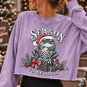 Puede incluir: Una sudadera corta de manga larga en color morado claro con el gr&aacute;fico de una rana con gorro de Pap&aacute; Noel y bufanda, sosteniendo una taza de cacao. El texto 'TIS THE SEASON TO BE JOLLY' rodea a la rana sentada sobre cajas de regalo.