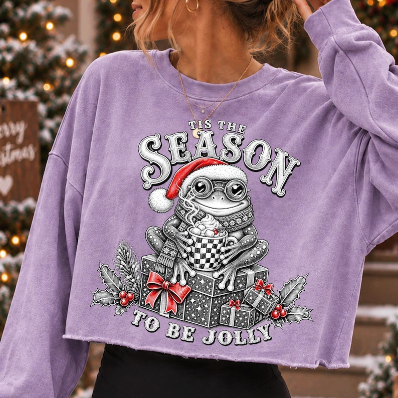 Puede incluir: Una sudadera corta de manga larga en color morado claro con el gr&aacute;fico de una rana con gorro de Pap&aacute; Noel y bufanda, sosteniendo una taza de cacao. El texto 'TIS THE SEASON TO BE JOLLY' rodea a la rana sentada sobre cajas de regalo.