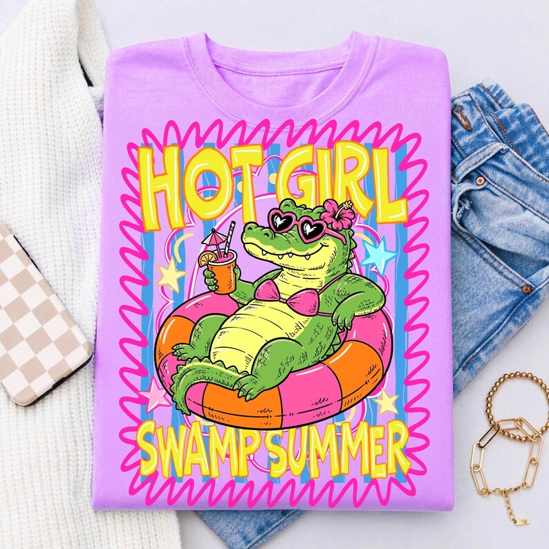 May include: A lilac t-shirt featuring a cartoon alligator in a pink bikini and heart-shaped sunglasses, relaxing on a pink and orange inner tube. The shirt has the words "HOT GIRL SWAMP SUMMER" in yellow and pink.